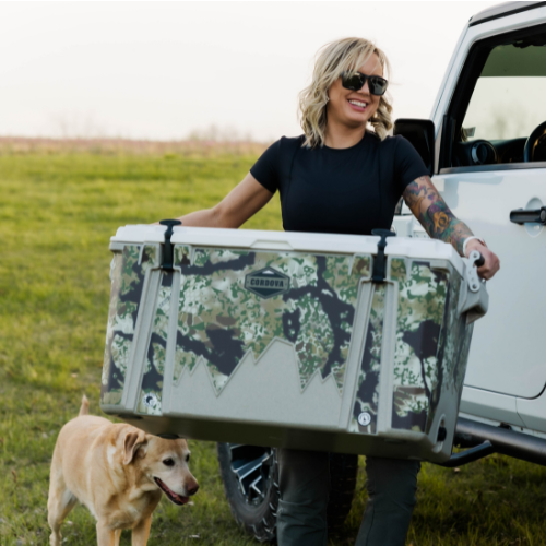 An outdoorsy woman carrying a large Kryptek Obskura camo hard cooler [88 qt] [48 qt]