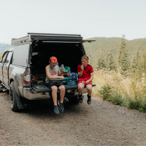Two boys sitting on a truck bed outside, eating their lunch from their insulated cooler lunch bags [Yonder]